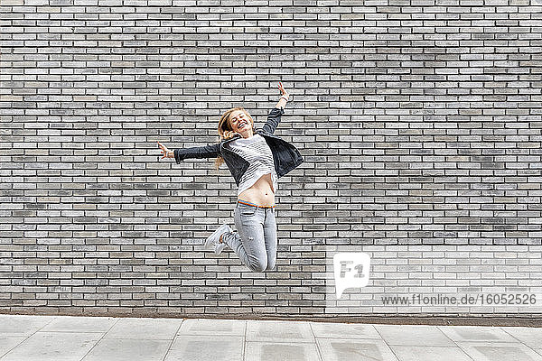 Carefree woman with arms outstretched jumping on footpath against gray brick wall
