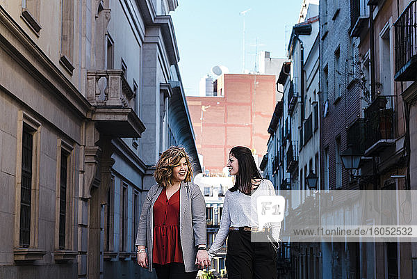 Lesbisches Paar hält sich an den Händen  während es inmitten von Gebäuden in der Stadt spazieren geht