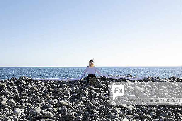 Frau mit künstlichen langen Händen sitzt auf Kieselsteinen am Strand gegen klaren Himmel