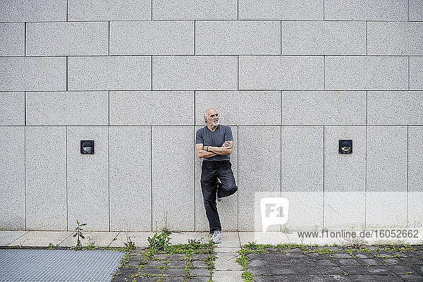 Thoughtful senior man with arms crossed leaning on wall in city