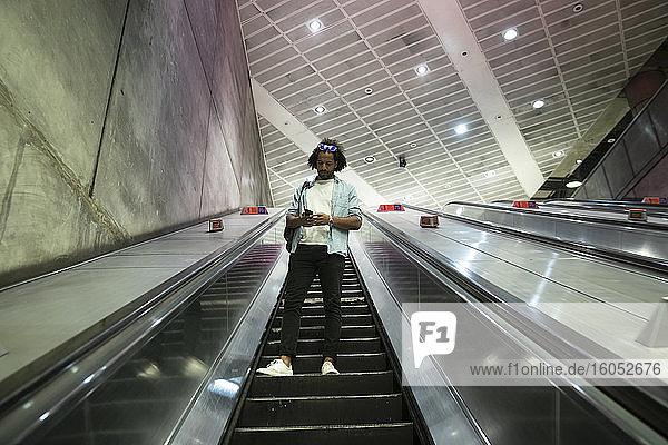 Junger trendiger Mann benutzt Smartphone auf Rolltreppe in U-Bahn-Station