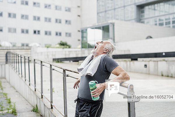 Müder älterer Mann  der eine Flasche hält und gähnt  während er an einem Geländer in der Stadt steht