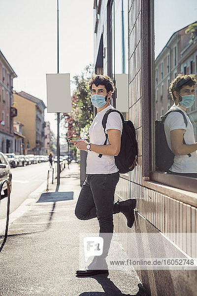 Mann mit Maske  der ein Smartphone benutzt  während er an einem Gebäude in der Stadt steht