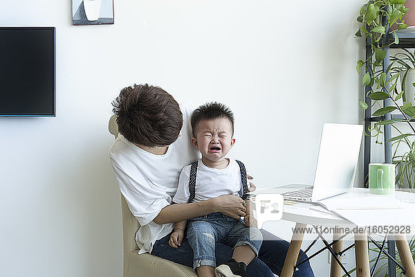 Mutter hält weinenden Jungen auf dem Schoß  während sie über einem Stuhl zu Hause sitzt