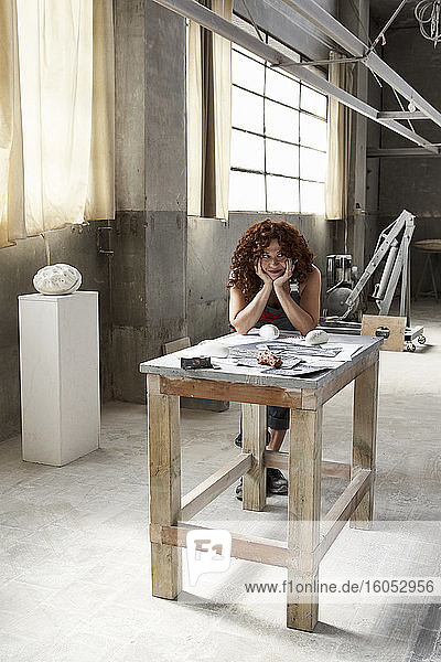 Steinmetzin mit Händen am Kinn  die sich mit Skizzen und Werkzeugen über eine Werkbank im Atelier lehnt