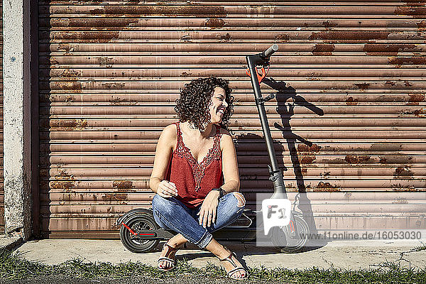 Smiling woman with curly hair sitting on electric push scooter against closed shutter
