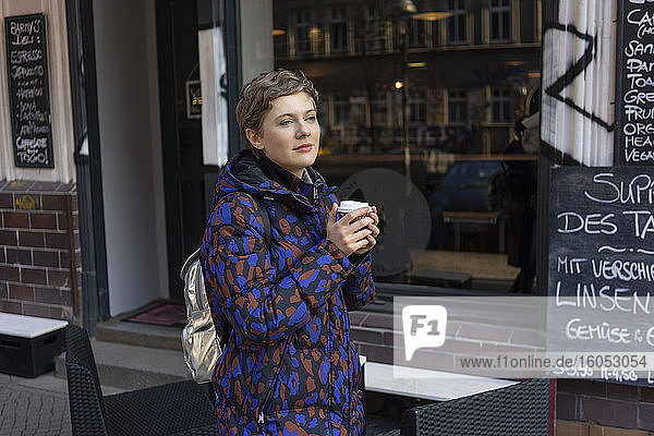 Porträt einer träumenden Frau mit Rucksack und Kaffee zum Mitnehmen vor einem Coffeeshop
