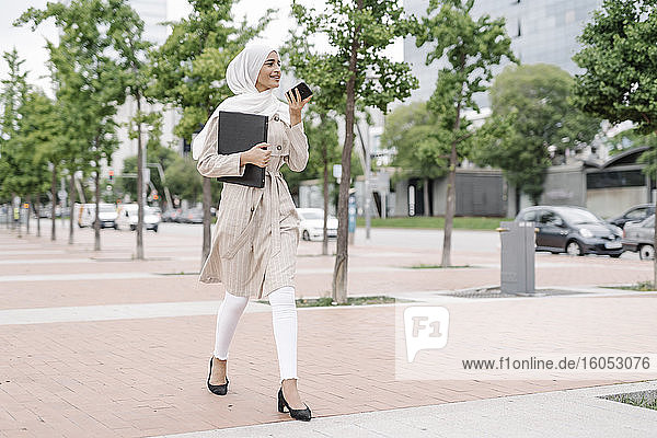 Muslimisches Mädchen im Teenageralter benutzt ein Smartphone und hält eine Mappe auf dem Bürgersteig in der Stadt