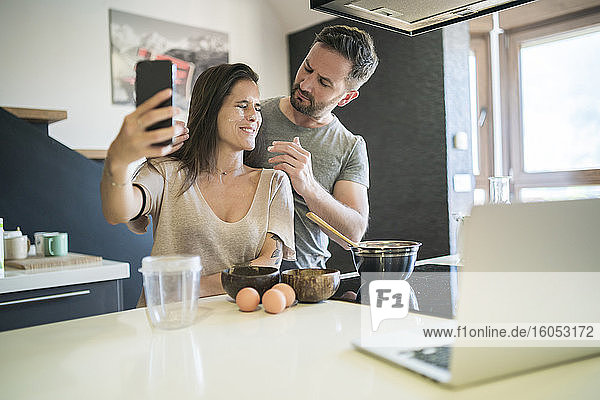 Verspielter Mann  der Mehl auf das Gesicht einer Frau aufträgt  während sie ein Selfie mit einem Smartphone zu Hause macht