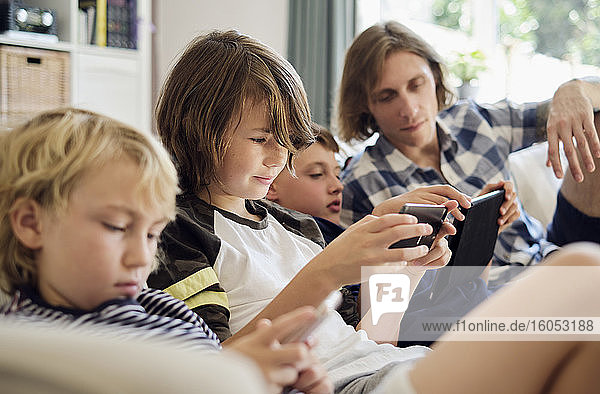 Father looking at boys playing games on smart phones and digital tablet in living room