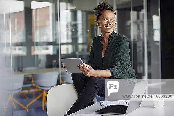 Porträt einer lächelnden Geschäftsfrau mit digitalem Tablet  die auf einem Schreibtisch sitzt und in die Ferne schaut