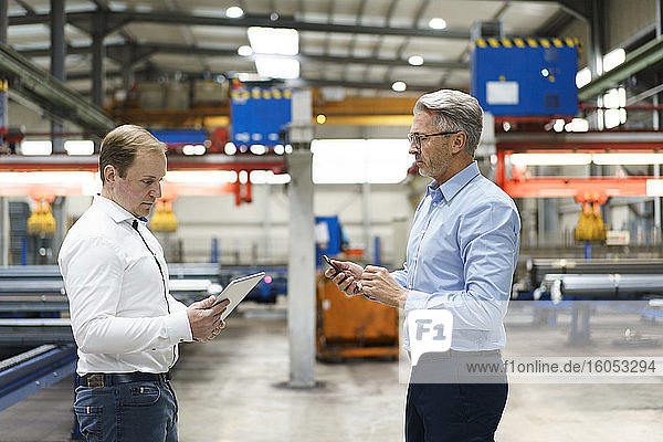 Zwei Geschäftsleute mit Tablet und Mobiltelefon bei einem Treffen in einer Fabrik