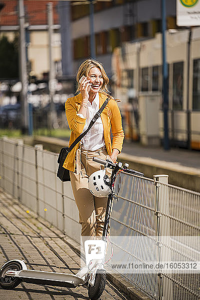 Fröhliche Geschäftsfrau  die ein Smartphone benutzt  während sie mit einem Roller auf dem Bürgersteig in der Stadt steht