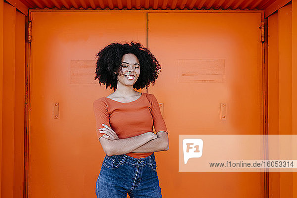 Lächelnde junge Afro-Frau steht mit verschränkten Armen vor einer orangefarbenen Tür