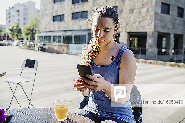 Sporty young woman using smart phone while sitting at sidewalk cafe in city