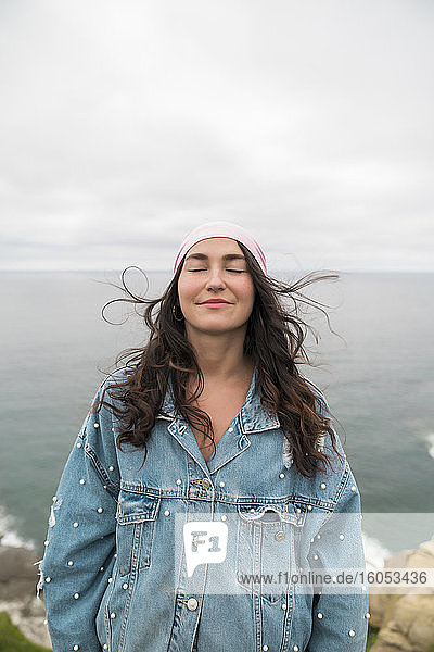 Lächelnde weibliche Krebsüberlebende mit geschlossenen Augen  die am Meer steht