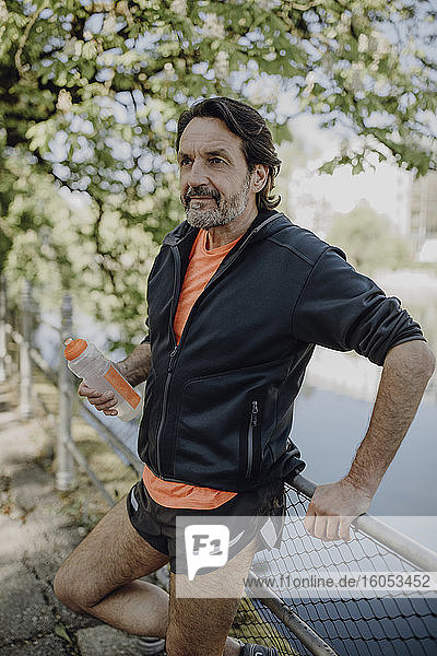 Nachdenklicher reifer Mann  der eine Wasserflasche hält  während er an einem See im Park steht