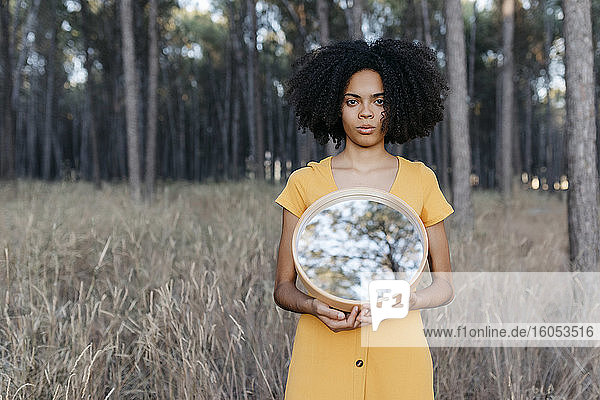 Junge Frau mit Afro-Haar  die einen Spiegel hält  während sie auf dem Land im Wald steht