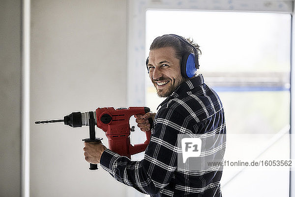 Porträt eines lächelnden Arbeiters  der eine elektrische Bohrmaschine auf einer Baustelle benutzt