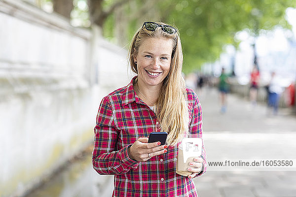 Lächelnde Frau mit Kaffee in der Hand  die ein Mobiltelefon benutzt  während sie auf dem Fußweg steht