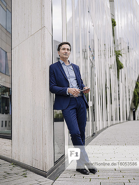 Serious mature businessman with tablet leaning against a building in the city