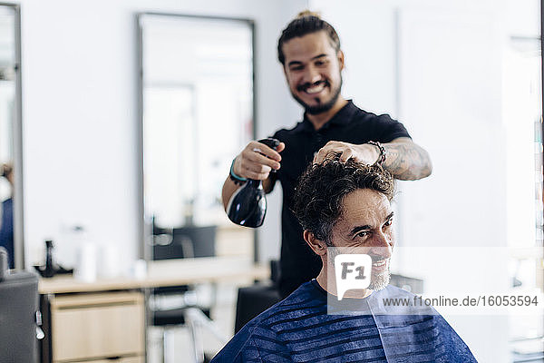 Fröhlicher Friseur sprüht Wasser auf das Haar eines Mannes im Salon