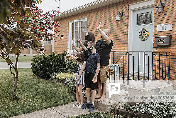 Eine Familie mit Masken winkt mit den Händen  während sie vor dem Haus im Hof steht