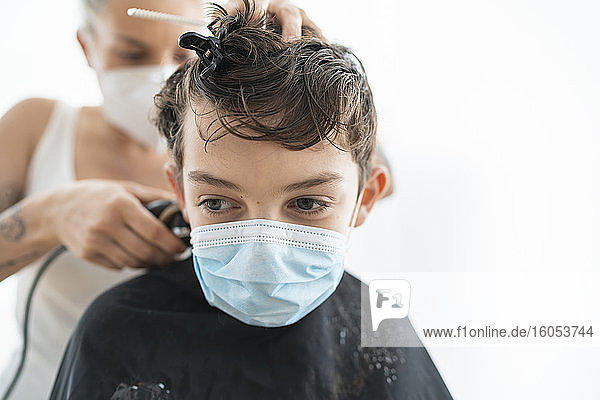 Nahaufnahme eines Friseurs  der einem Jungen mit Maske zu Hause während der Ausgangssperre die Haare schneidet