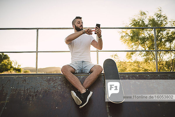 Gut aussehender junger Mann  der sein Smartphone benutzt  während er auf einer Sportrampe im Skateboardpark sitzt