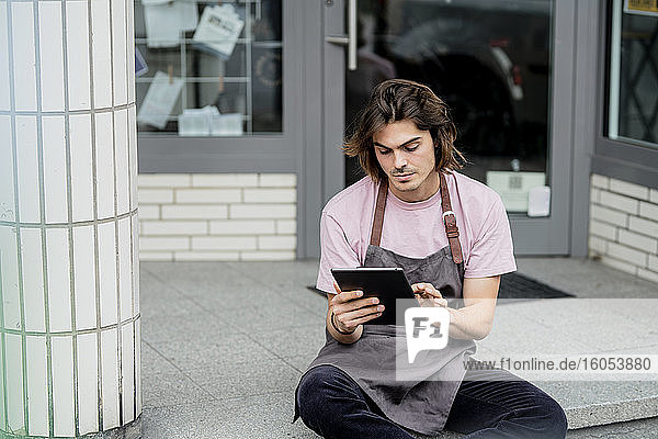 Male barista using digital tablet while sitting outside coffee shop