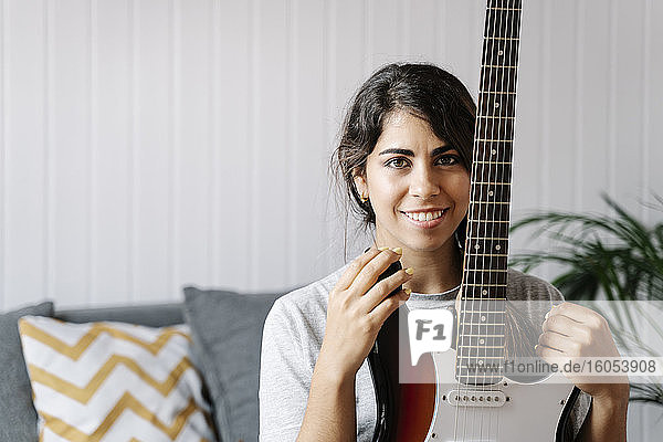 Selbstbewusste junge Frau  die eine E-Gitarre hält  während sie zu Hause auf dem Sofa sitzt