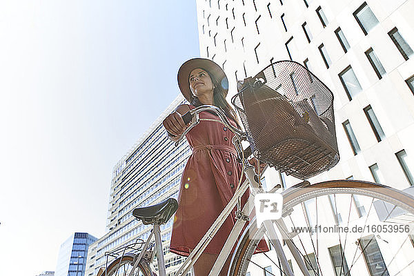 Nachdenkliche Frau steht mit Fahrrad gegen Gebäude und Himmel