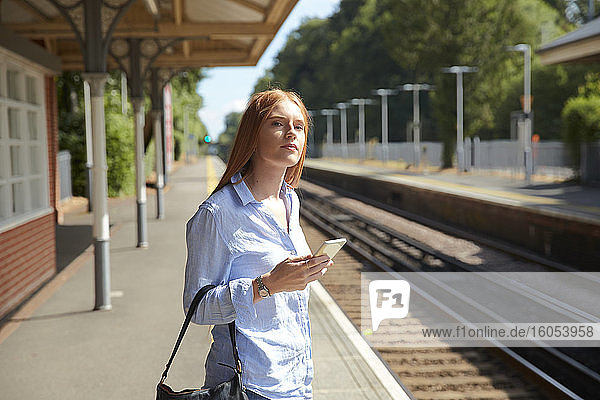 Junge Frau hält ihr Smartphone auf dem Bahnsteig eines Bahnhofs