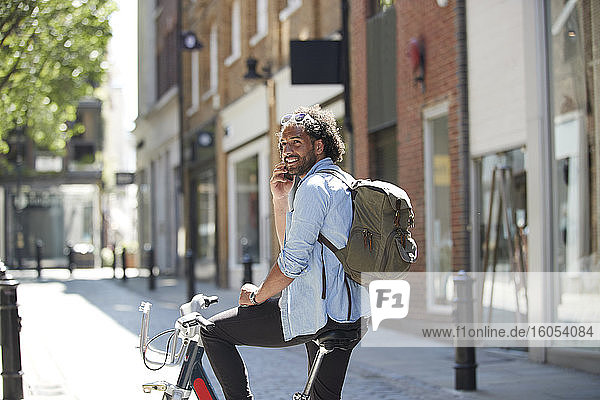Porträt eines lächelnden jungen Mannes am Telefon mit Leihfahrrad und Rucksack in der Stadt  London  UK