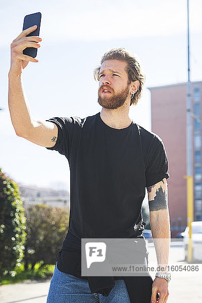 Bärtiger Mann  der ein Selfie macht  während er an einem sonnigen Tag in der Stadt vor einem klaren Himmel steht