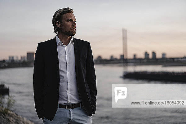 Businessman standing at the riverside at dusk looking away