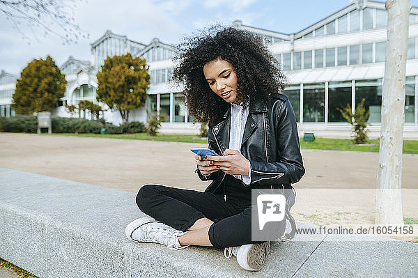 Young afro woman using smart phone while sitting cross-legged on retaining wall