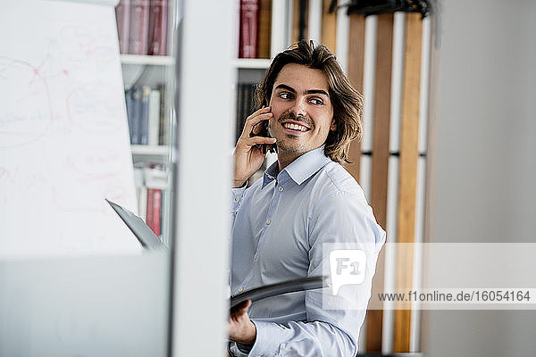 Lächelnder Geschäftsmann  der über sein Smartphone spricht und wegschaut  während er im Büro sitzt