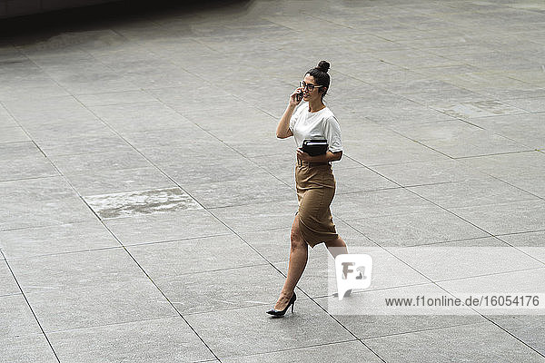 Junge Geschäftsfrau  die beim Gehen auf einem Fußweg in der Stadt mit ihrem Mobiltelefon spricht
