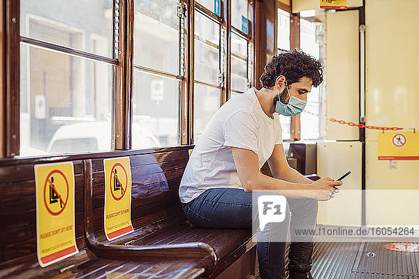 Mann mit Gesichtsmaske benutzt Mobiltelefon in der Straßenbahn