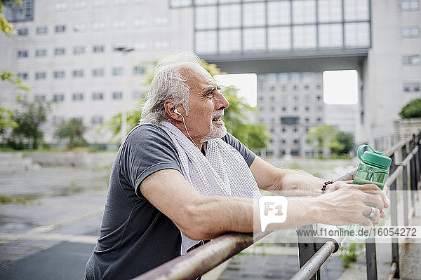 Älterer Mann hält Wasserflasche und hört Musik  während er am Geländer steht