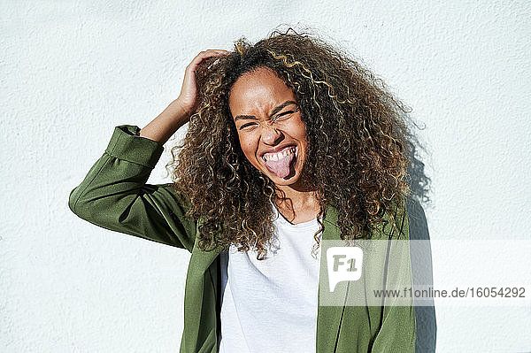 Verspielte Frau streckt ihre Zunge gegen eine weiße Wand an einem sonnigen Tag aus
