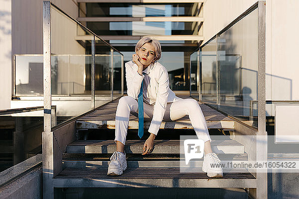Junge Frau im weißen Anzug sitzt auf den Stufen eines modernen Gebäudes