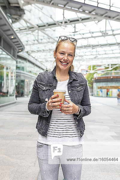 Lächelnde erwachsene Frau  die eine Kaffeetasse hält  während sie auf dem Gehweg in der Stadt steht