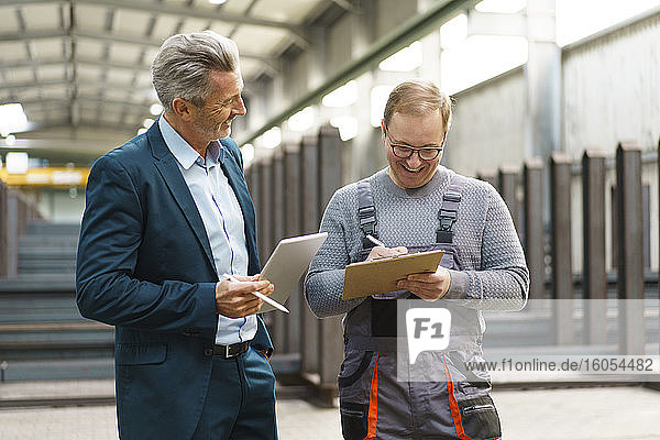 Happy businessman with tablet and employee with clipboard in a factory
