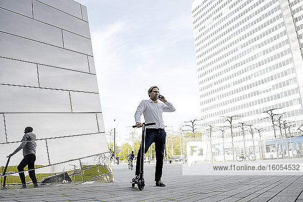 Geschäftsmann mit Kick-Scooter und Smartphone spiegelt sich in Gebäudefassade