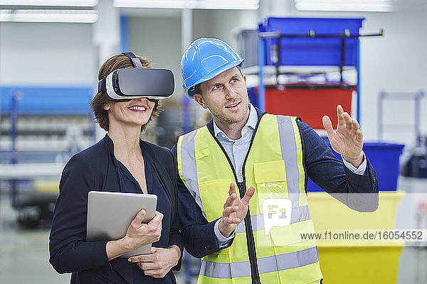 Ein Vorgesetzter erklärt einer weiblichen Führungskraft  die eine VR-Brille trägt und ein digitales Tablet in einer Fabrik hält  einen Plan