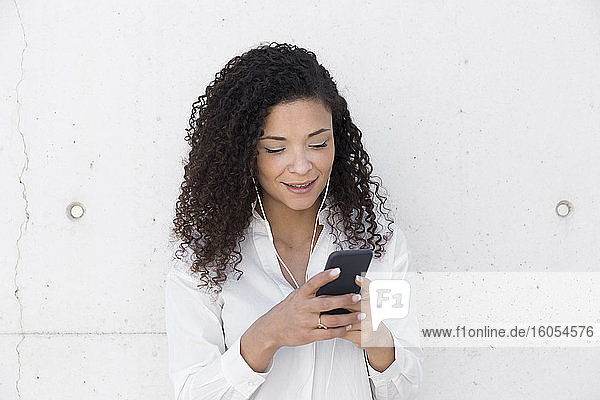 Geschäftsfrau mit lockigem Haar  die ein Smartphone benutzt  während sie an der Wand steht