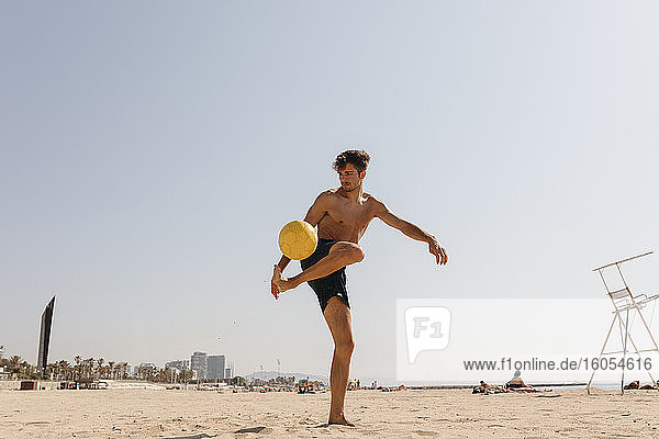 Hemdloser männlicher Sportler spielt mit Ball am Strand gegen klaren Himmel an einem sonnigen Tag