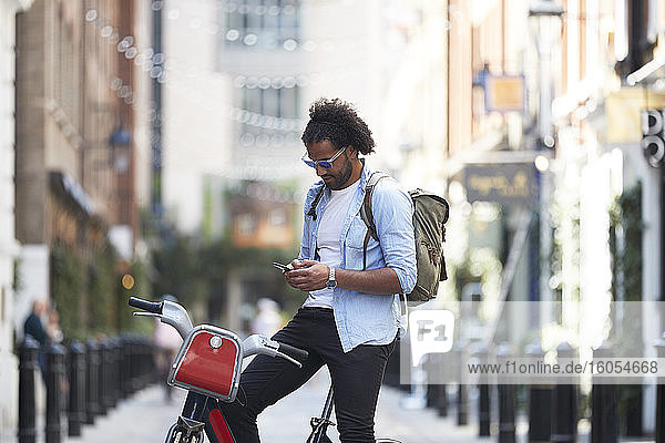 Junger Mann mit Leihfahrrad und Rucksack  der sein Handy in der Stadt benutzt  London  Großbritannien